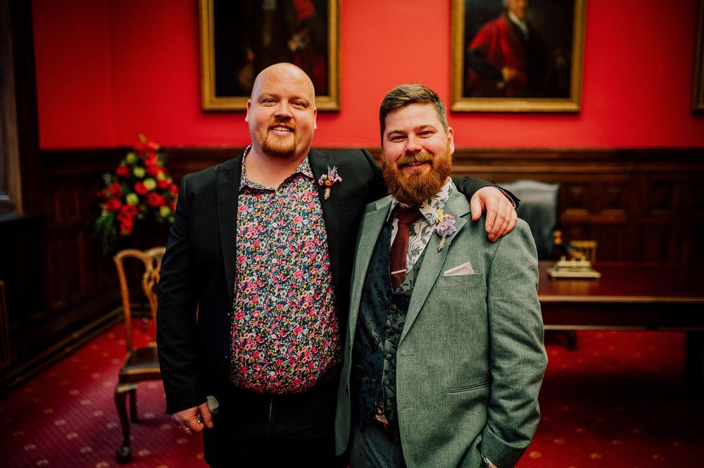 Two people in formal attire pose together in a red room with framed portraits on the wall.