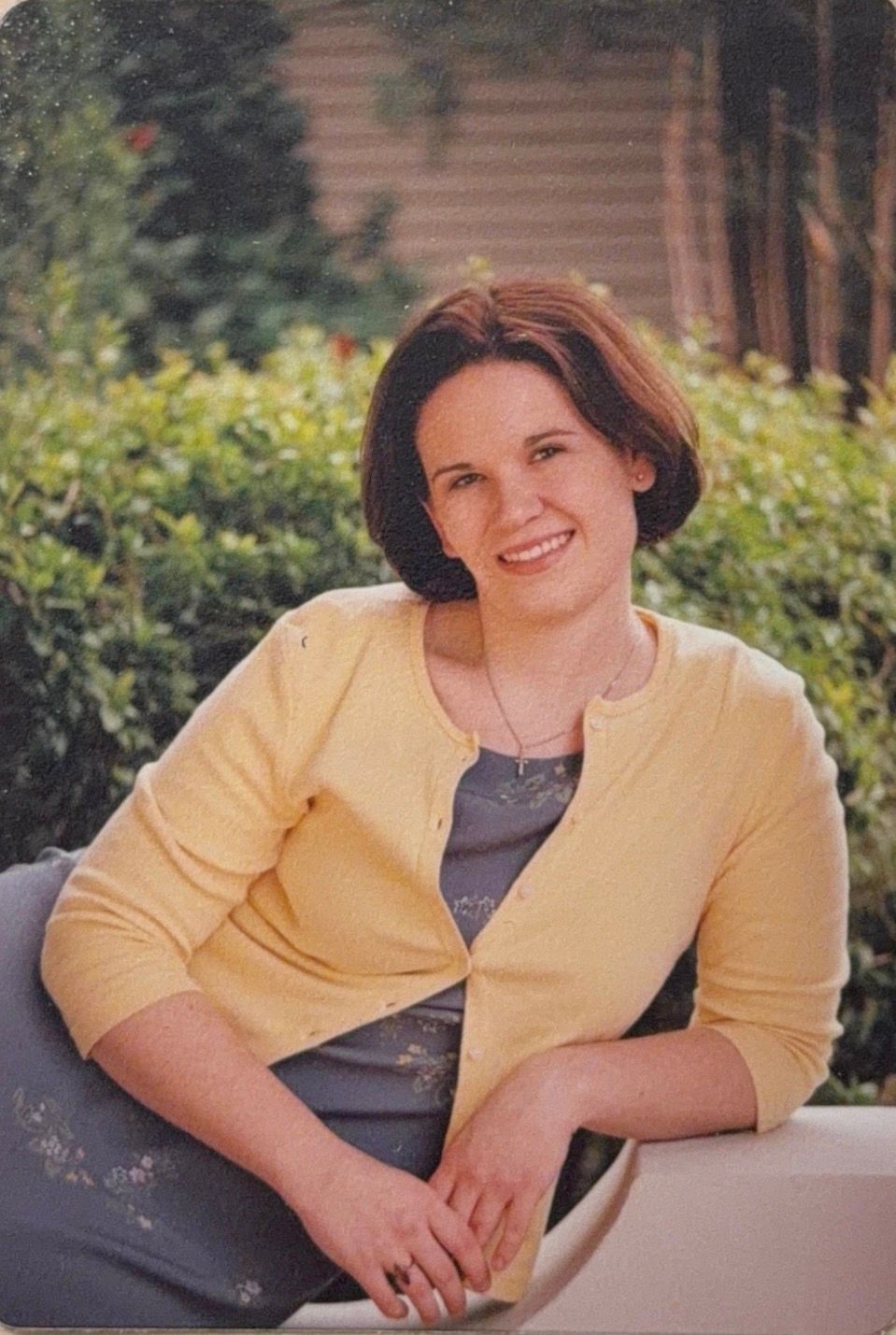 A person in a yellow button-up shirt and dark top sits casually on an outdoor patio in natural lighting.