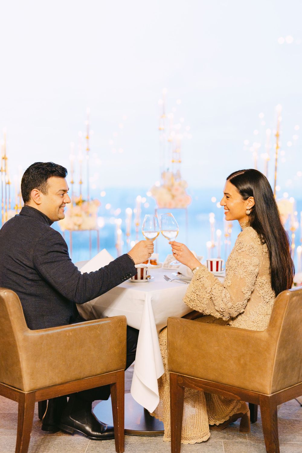 Couple toasting champagne glasses at an elegant restaurant with scenic harbor views.