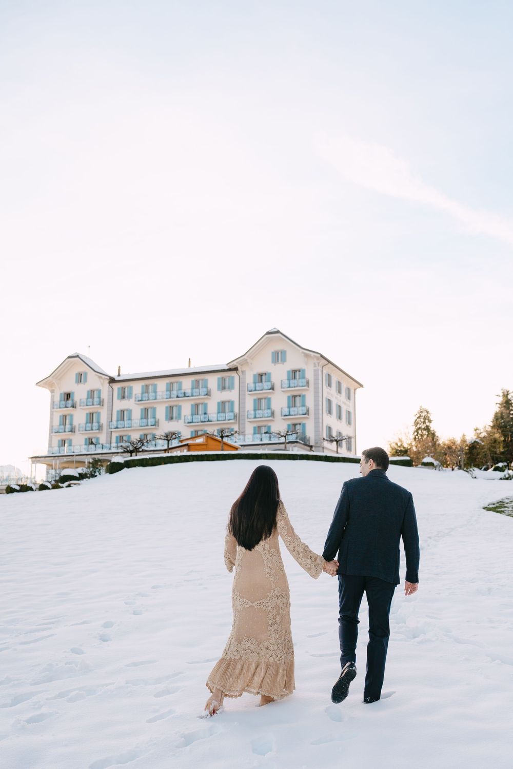 A couple walks hand in hand through a snowy landscape with a grand white hotel building visible in the background.