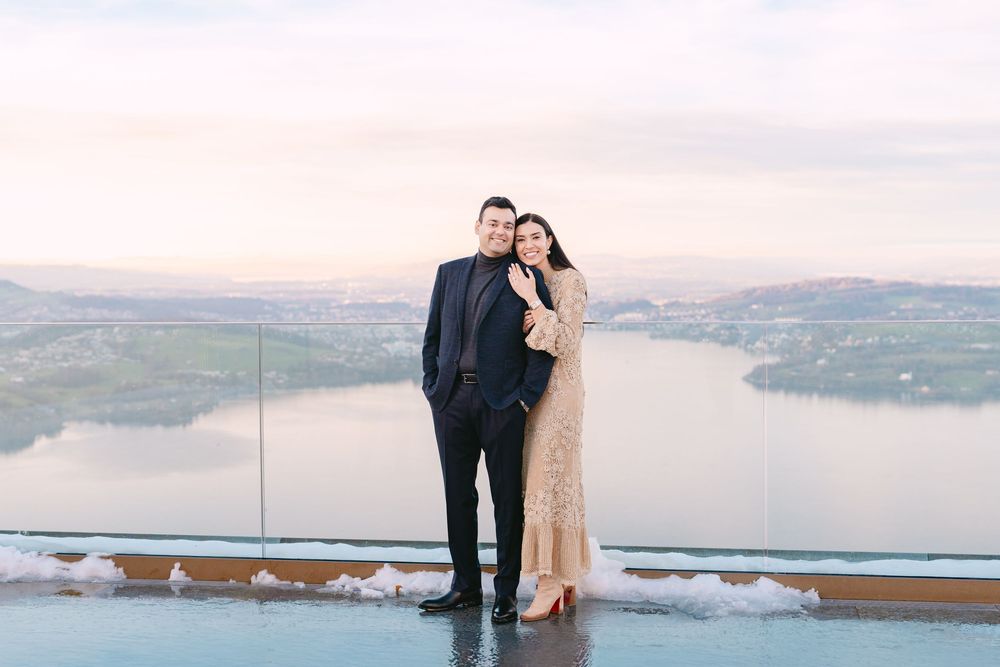A couple stands together on a scenic overlook during sunset with a lake and mountains in the background.
