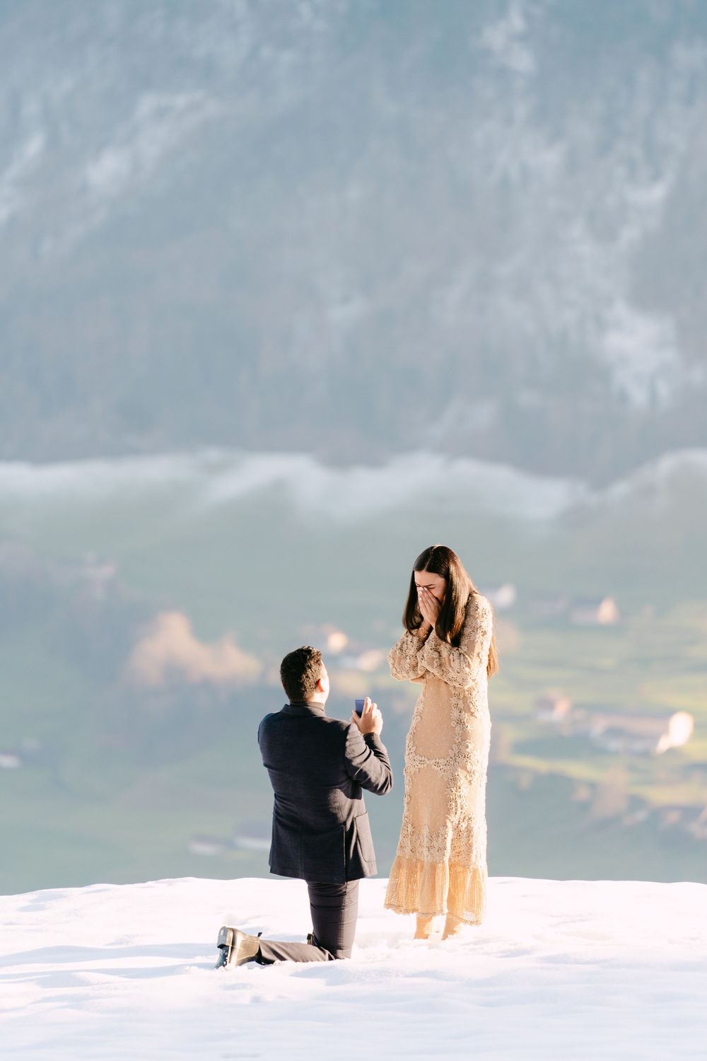 A romantic proposal scene on a snowy mountain peak with dramatic cloudy skies in the background.