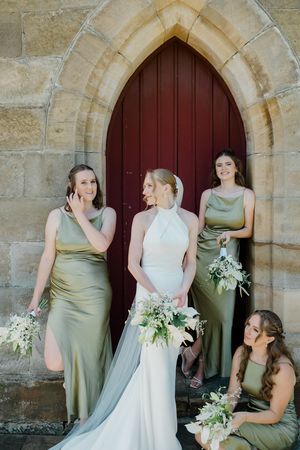 A wedding party in sage green bridesmaid dresses and white bridal gown pose together by an arched church doorway.