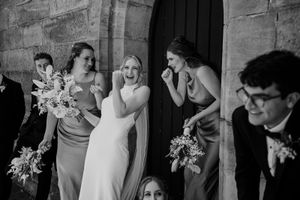 A candid black and white wedding party moment captured at a church entrance with bouquets and formal attire.