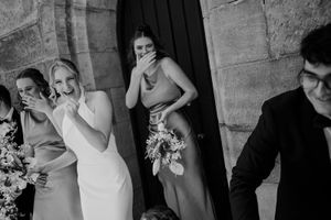 Black and white photo of wedding party members sharing a lighthearted moment outside a stone church entrance.
