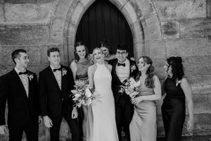 Wedding party members in formal attire pose together in front of a stone building with pink and white bouquets.