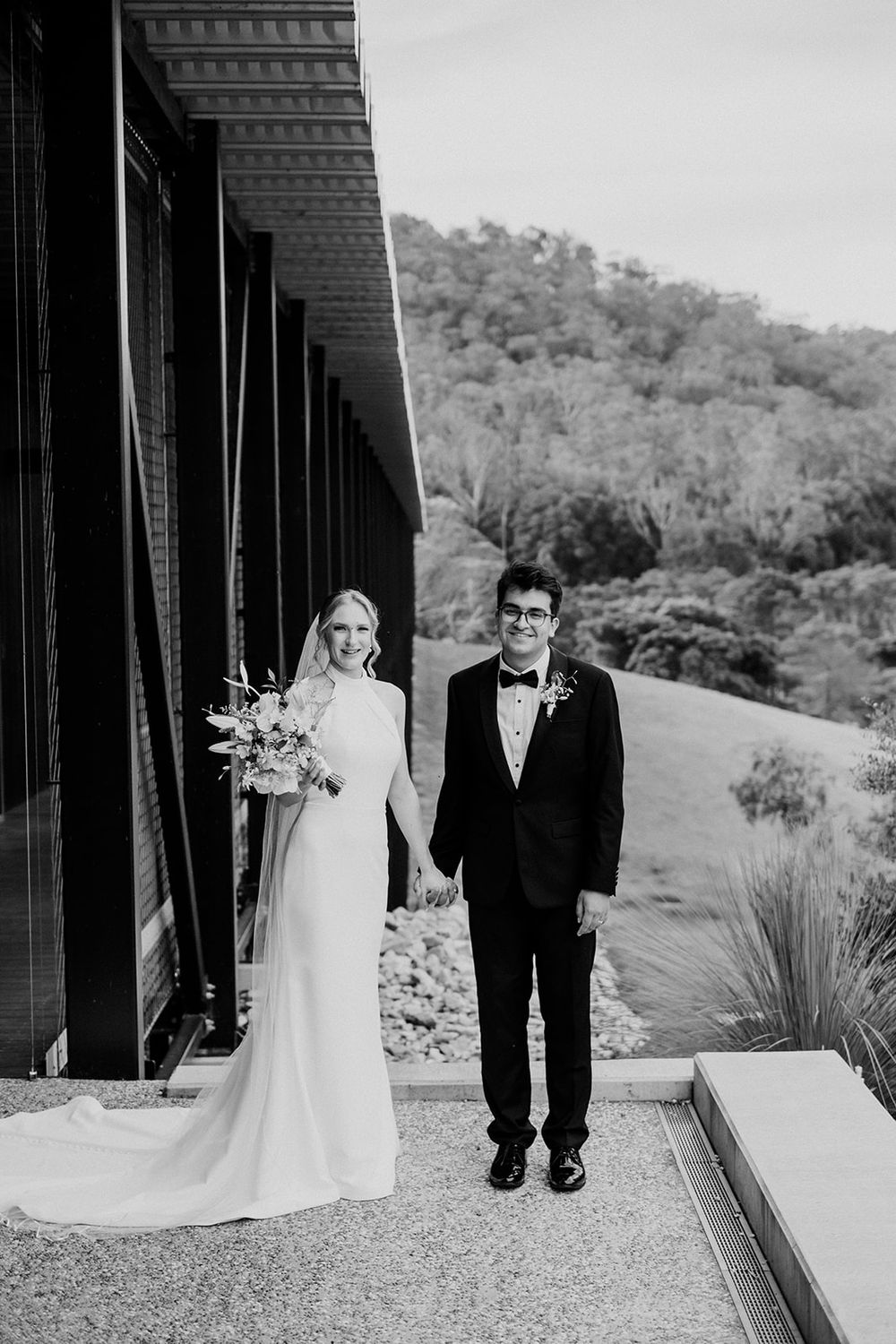 Black and white wedding photo showing couple standing together on patio with scenic hillside backdrop.