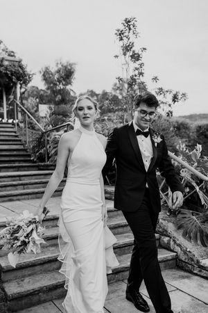 A couple in wedding attire walks down stone garden steps in a black and white photograph.