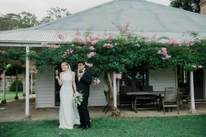A wedding party walks across a lush green lawn in front of a white country house with wraparound porch.