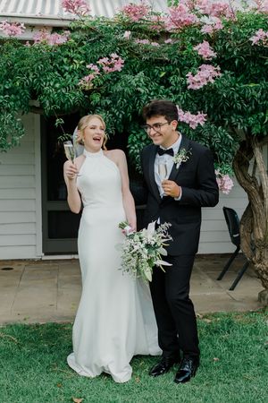 A couple in wedding attire shares a joyful moment under a blooming pink floral tree in a garden setting.