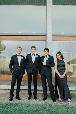 Wedding party in formal black attire poses together against modern building exterior.