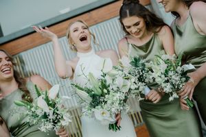 Wedding party in sage green dresses laughing together while holding white floral bouquets.