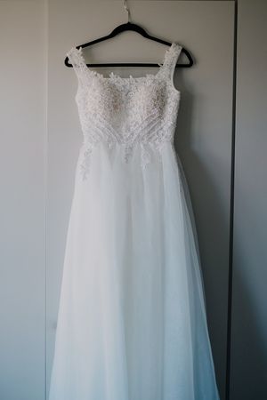 White wedding dress with lace bodice and flowing tulle skirt hangs on black hanger against light blue wall.