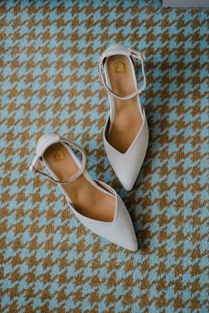 Elegant white pointed-toe bridal flats with ankle straps displayed on a blue and tan houndstooth patterned background.