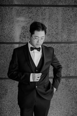 Black and white portrait of someone in a classic tuxedo with bow tie standing against a textured wall.