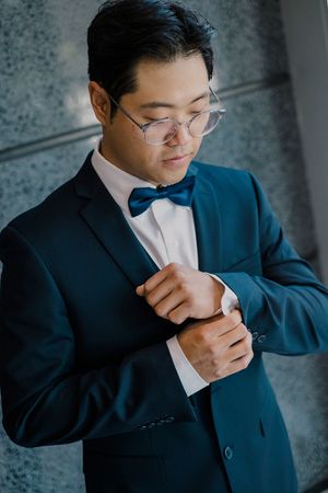 A person in a navy suit and bow tie adjusts their cuff while wearing glasses in a formal portrait setting.
