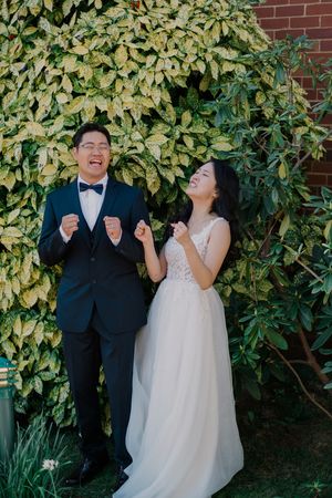 A joyful couple in wedding attire share a candid moment while standing against a leafy green and yellow backdrop.