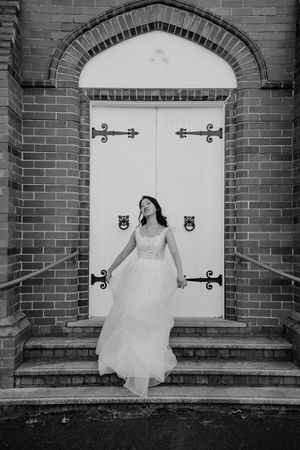 A bride in a flowing white dress stands in front of an elegant white church door with decorative black hinges.