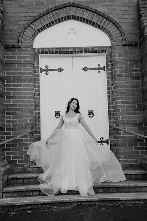 A black and white photo of an elegant wedding dress flowing in front of a white arched doorway with ornate brick details.