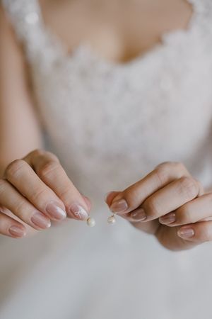 Close-up of manicured hands with nude polish holding small pearl beads against white lace background.