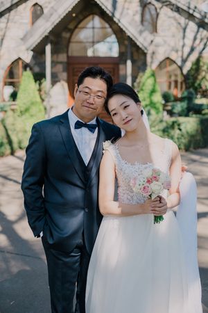 A wedding couple poses together outdoors in formal attire under stone archways on a sunny day.
