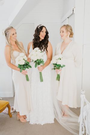 Bridesmaids in white dresses holding white rose bouquets share candid moments during a bright indoor wedding photoshoot.