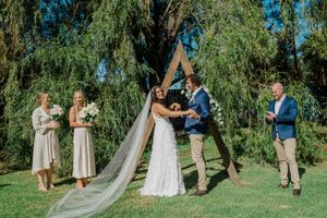 Outdoor wedding ceremony under a wooden arch with willow trees in the background on a sunny day.