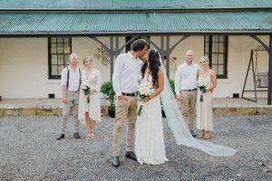 Wedding party poses in front of rustic blue-roofed building during outdoor countryside celebration.