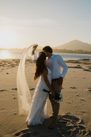 A couple in wedding attire shares an intimate moment on a sandy beach at sunset with mountains in the background.