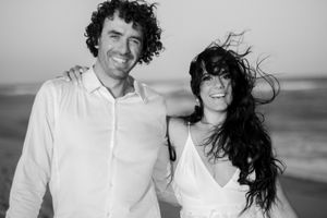 A couple shares romantic moments on a beach in black and white photos wearing white clothing with wind-swept hair.