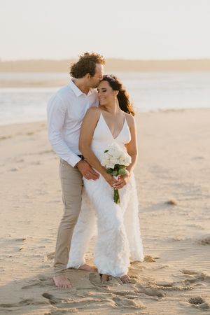 A romantic beach wedding couple embraces on sandy shores at sunset holding a white bouquet.
