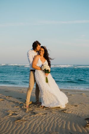 A couple in wedding attire shares romantic moments on a sandy beach with the ocean waves in the background.