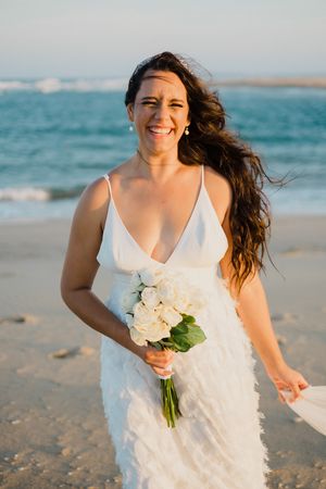 A person in a white wedding dress holds a bouquet of cream roses while walking joyfully on a sunny beach.