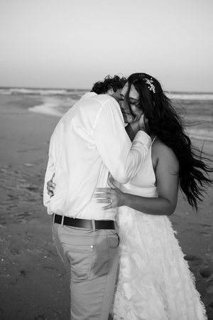 A romantic couple in white attire share an intimate moment on a beach during sunset in black and white photography.