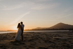 Silhouettes of a couple embrace on a beach at sunset with mountains in the background.