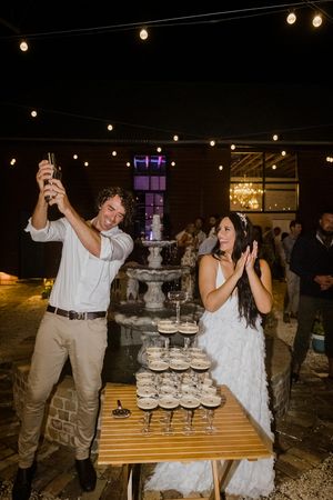A couple pours champagne over a tiered wedding cake display at an outdoor evening reception.