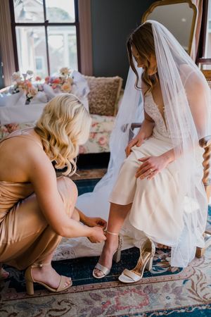 Bridesmaid helps bride put on her wedding shoes while getting ready in an elegant room.