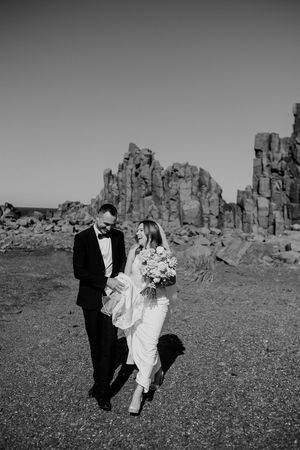 A couple in wedding attire walks together against dramatic rock formations in a black and white desert landscape.