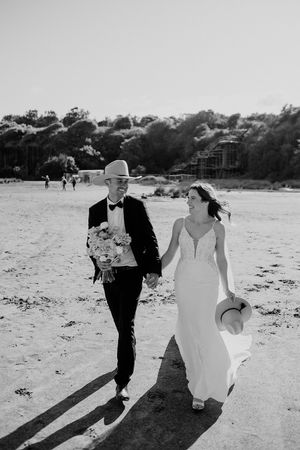 A couple in wedding attire walking together on a sunny beach, holding hands and carrying a bouquet and hat.