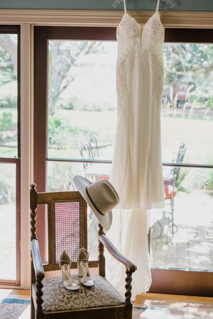 A white wedding dress hangs by a window with bridal shoes and a hat placed on an antique wooden chair.