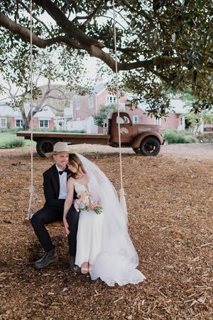 A couple sits on a rustic swing beneath a tree with a vintage truck in the background at their countryside wedding.