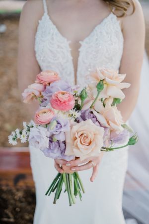 A delicate pastel bridal bouquet featuring pink ranunculus, peach roses, and purple blooms against a white lace dress.