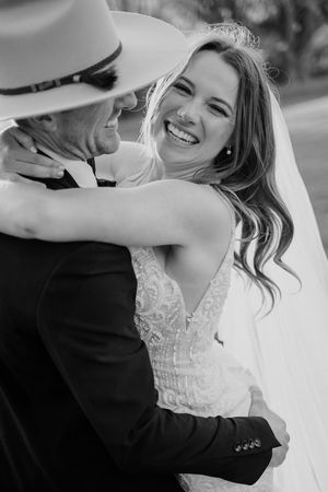 Black and white candid wedding photo showing joyful embrace between bride in lace dress and groom wearing hat.