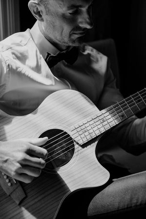 A musician in formal attire plays an acoustic guitar in dramatic black and white lighting.