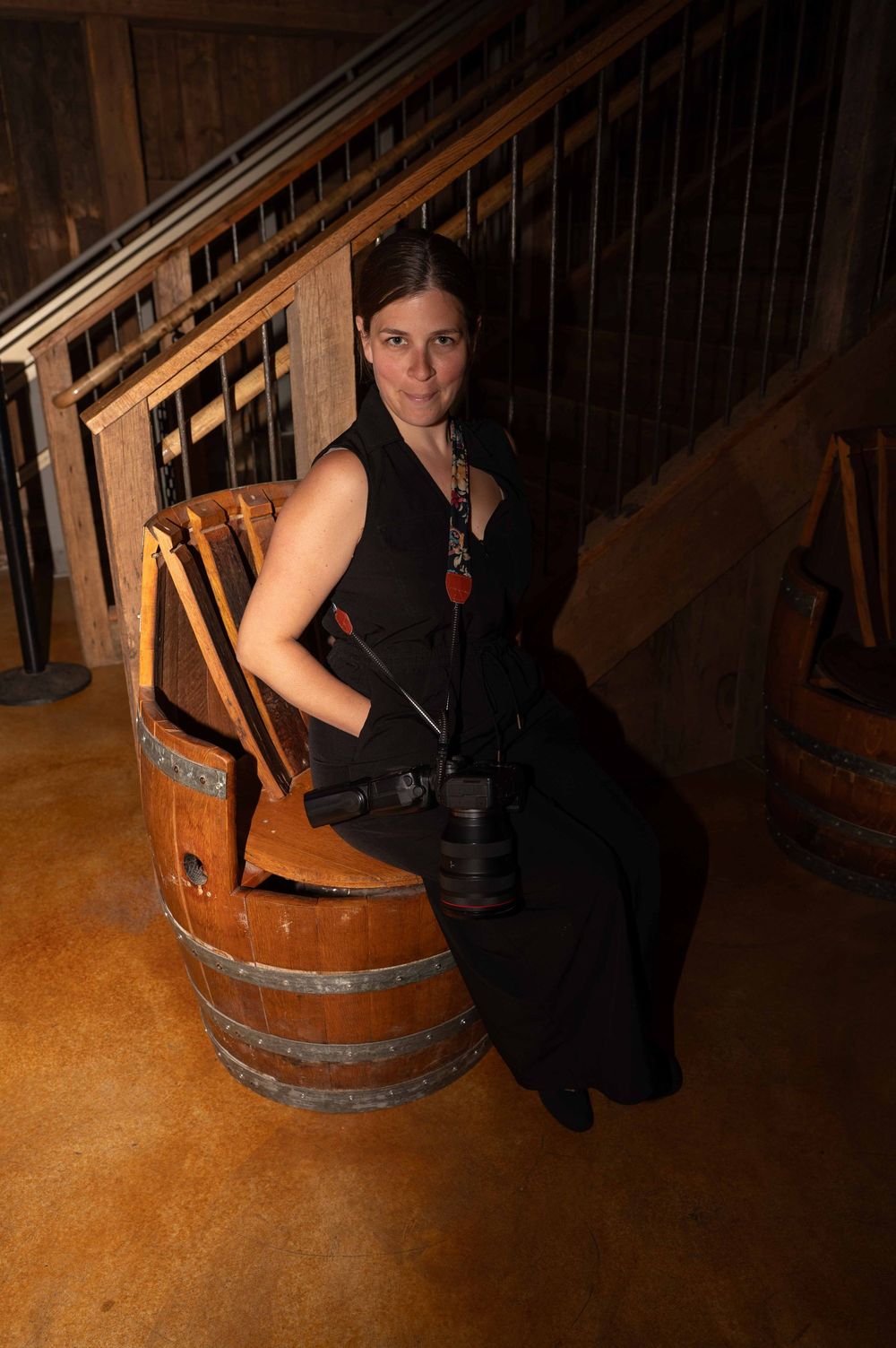 A person in a black dress poses on a wooden wine barrel in a rustic indoor setting.