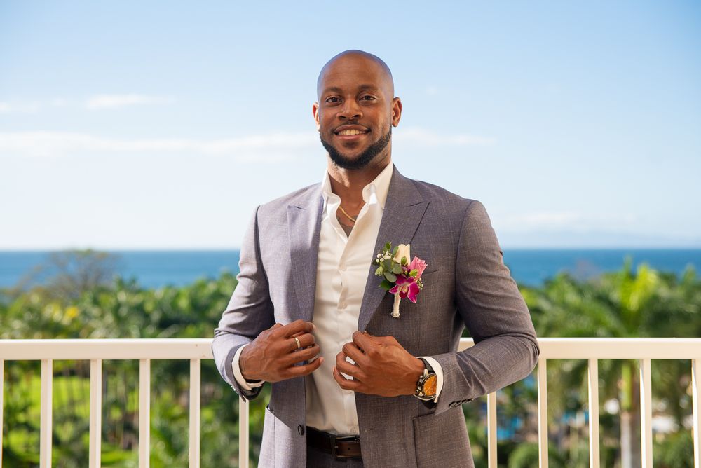 A groom in a grey suit adjusts his jacket while standing on a balcony with tropical scenery in the background.