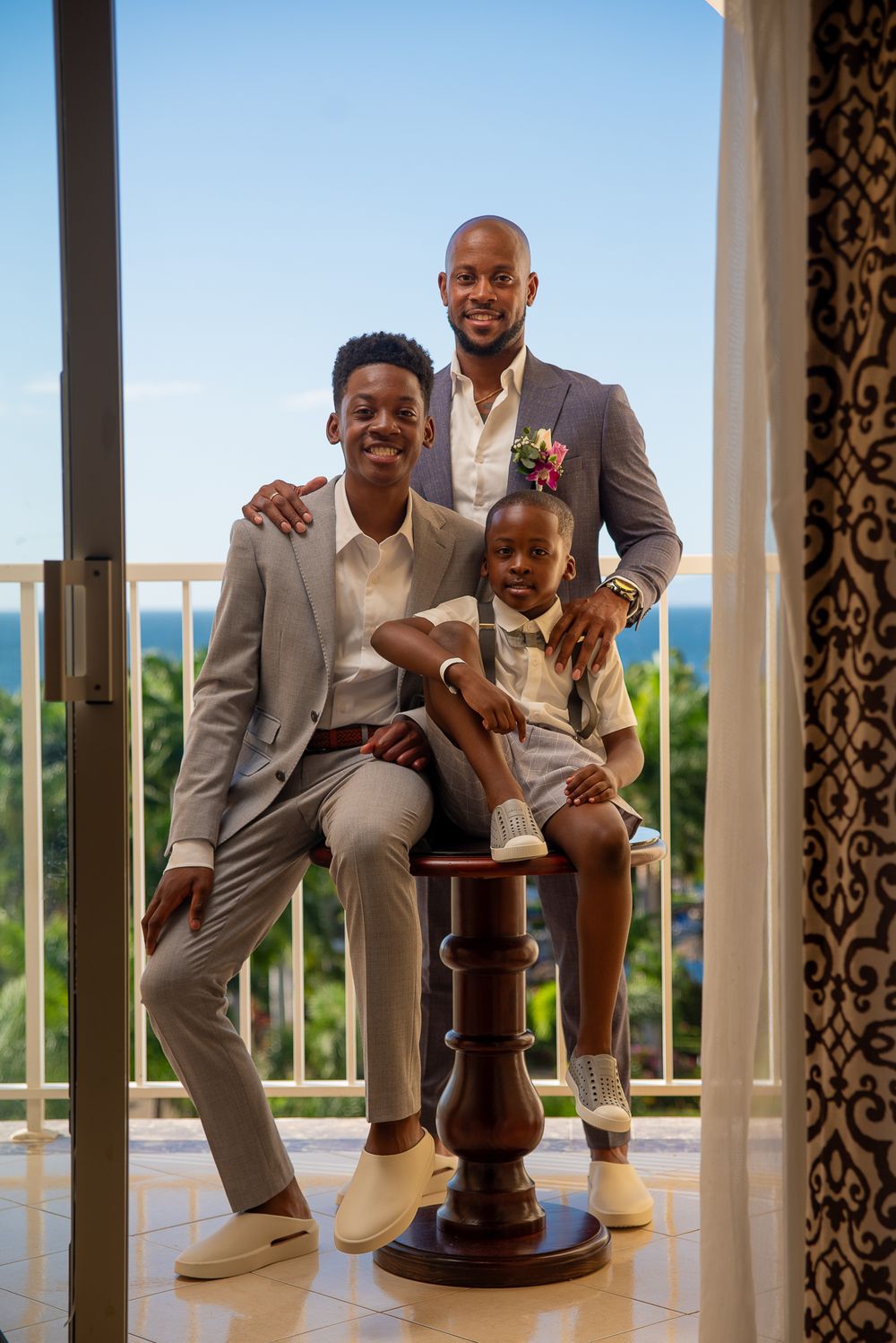 Three people in coordinated formal attire pose together on a balcony with an ocean view in the background.