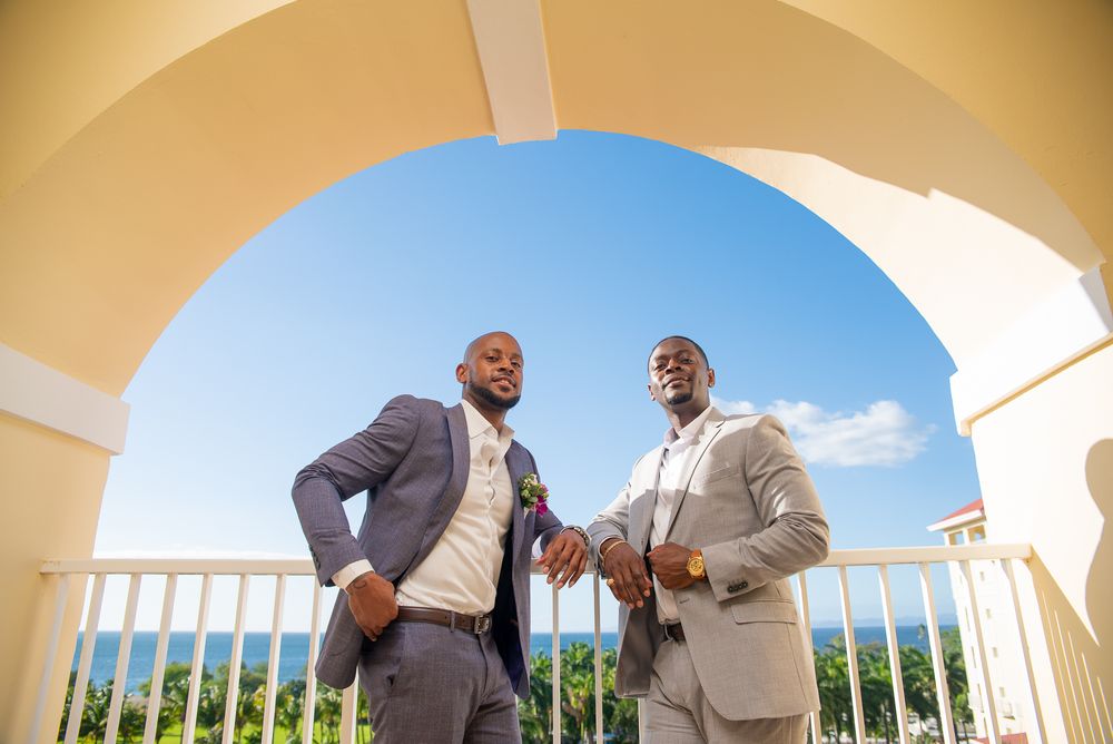 Groomsmen in gray suits share laughs and conversation on an outdoor porch with white columns and blue sky views.