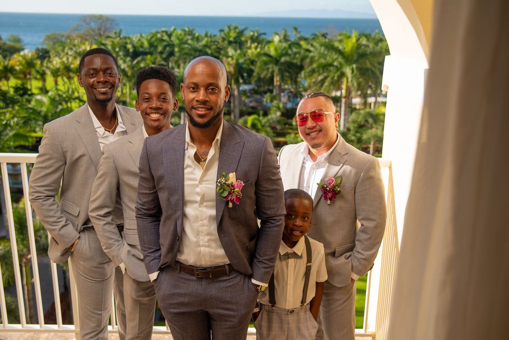 A group wearing matching light gray suits and white shirts pose together at an outdoor venue with greenery in background.