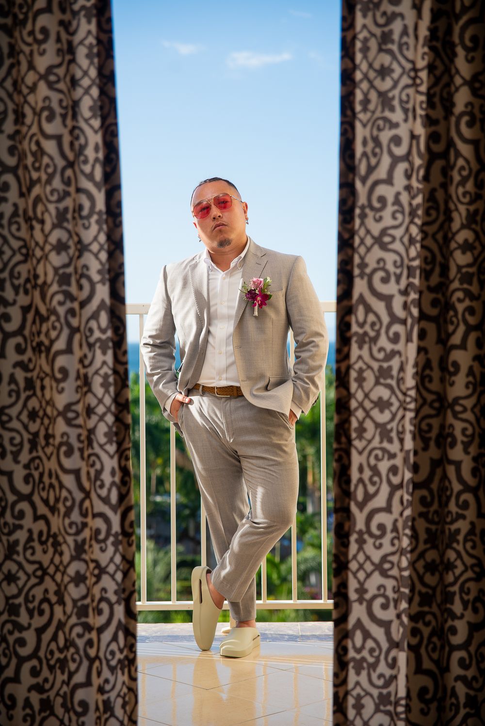 Stylish groom in light gray suit and pink boutonniere poses confidently between patterned curtains on balcony.